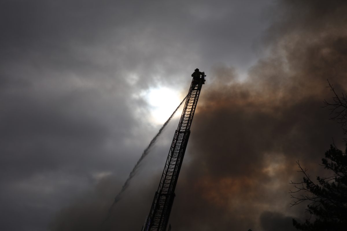 A firefighter operates a fire hose from atop a ladder as smoke plumes. 
