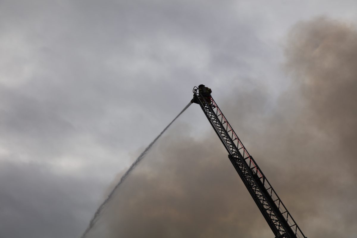 A firefighter operates a fire hose from atop a ladder as smoke plumes. 