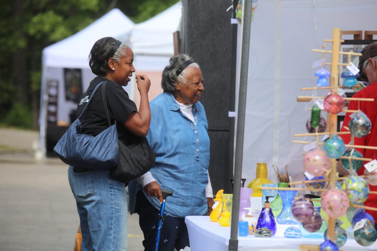 Two attendees of the Sunset Hills Makers Market talk to vendors at a booth selling glass art.