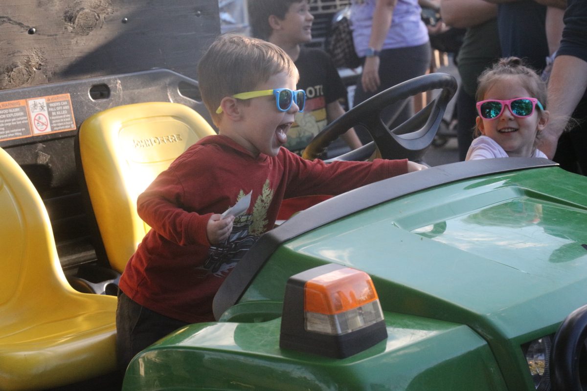 Following the lead of his companion, a young attendee gleefully honks the horn of the John Deere vehicle he sits in.